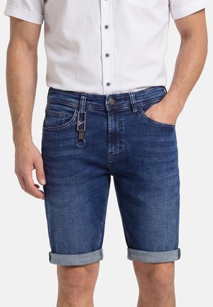 Mann trägt dunkelblaue, hochgekrempelte Jeansshorts mit angebrachtem Schlüsselanhänger und weißes, kurzärmliges Hemd mit Knöpfen, eine Hand in der Hosentasche.