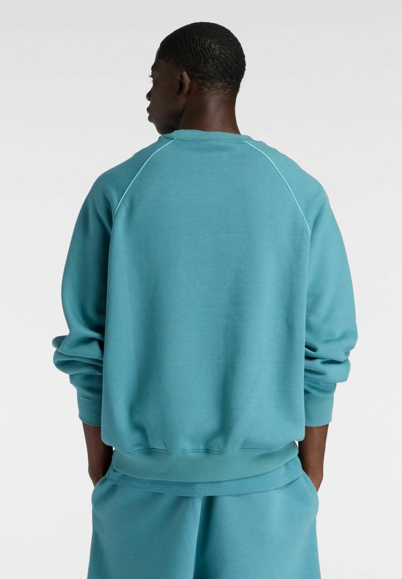 Uomo che indossa una felpa larga color teal e pantaloncini coordinati, in piedi con le mani nelle tasche, rivolto di spalle contro uno sfondo semplice.