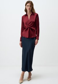 Blusa di raso color marrone scuro con un drappo frontale, maniche lunghe e scollatura a V, abbinata a una gonna fluente blu navy. Tacchi a punta in tonalità neutre.