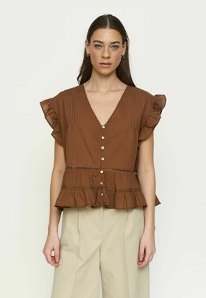 Žena má na sebe hnedú blúzku s volánikmi a krátkym rukávom s gombíkmi a krémové nohavice s širokými nohavicami, stojaca pred jednoduchým svetlým pozadím.