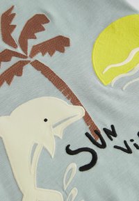 Broderet stof med en lyseblå baggrund, en hvid delfin, en brun palmetræ, en gul sol og sort tekst "Sol". Teksturering og stingdetaljer synlige.