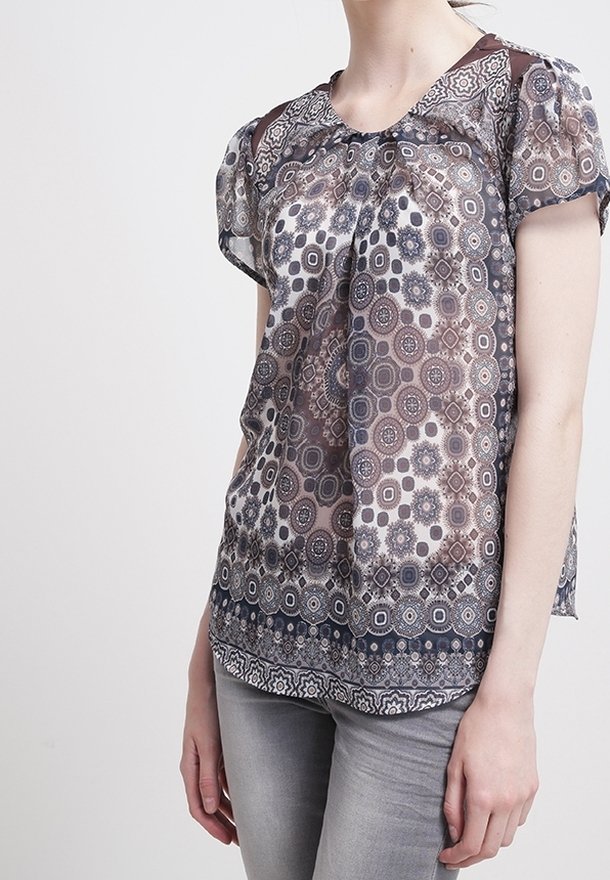 Femme portant un chemisier à manches courtes avec des motifs géométriques circulaires en marron, noir et blanc, associé à un pantalon gris clair.