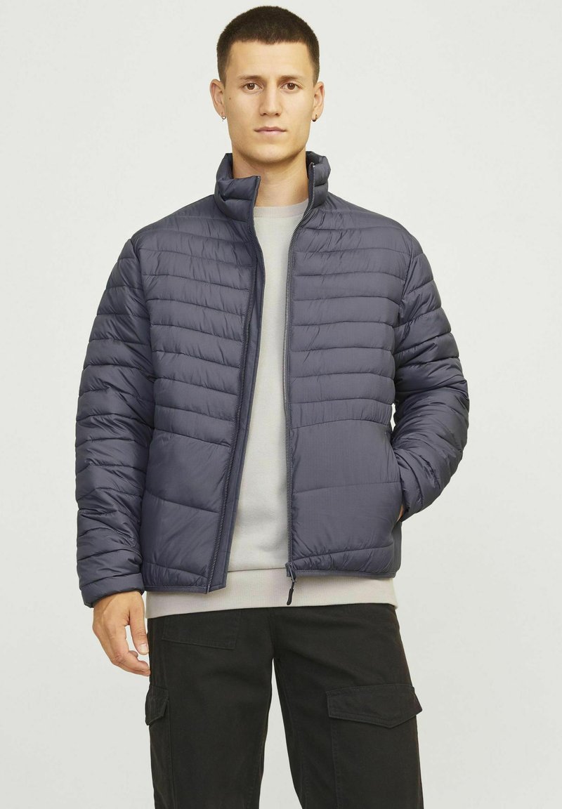Giacca imbottita di colore blu scuro, con zip frontale, colletto alto e cuciture orizzontali. Indossata su un maglione grigio chiaro.