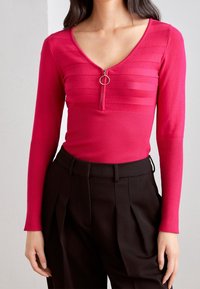 Maglietta aderente a maniche lunghe di colore rosa brillante, con zip a metà e una parte superiore circolare, texture a righe e sezione inferiore a coste.