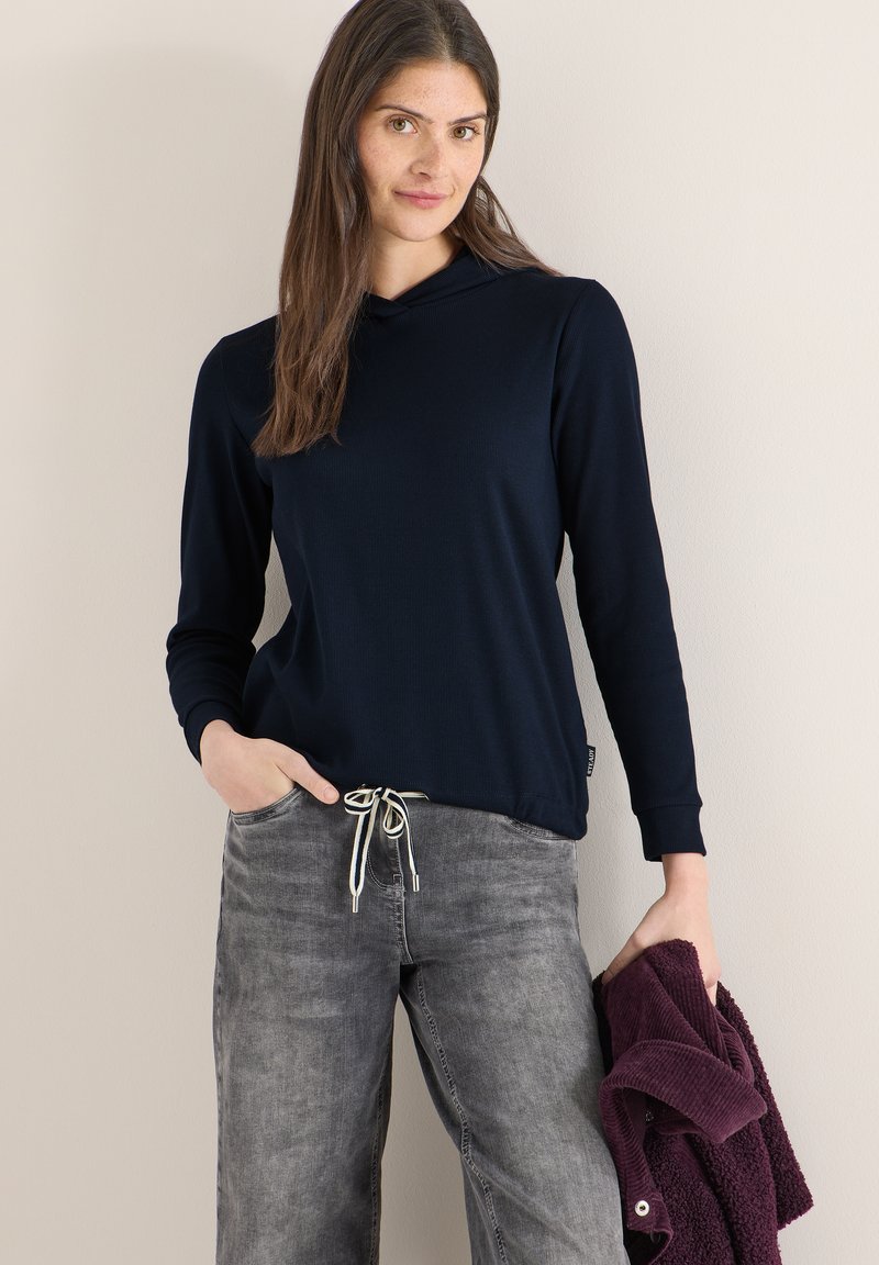 Femme portant un haut à manches longues bleu marine et un jean gris, tenant une veste pliée violet foncé dans une main, debout contre un mur beige.