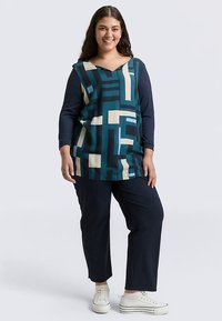 Top met lange mouwen met geometrisch patroon in blauw, groenblauw en beige; V-hals; gecombineerd met marineblauwe broek en witte sneakers.