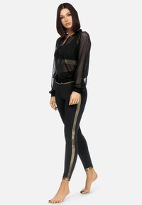 Czarna przezroczysta bomberka z siateczki z elastycznym dołem i mankietami, zestawiona z czarnymi legginsami z złotymi paskami po bokach i akcentami na kostkach.