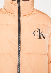 Puffer jacket in light peach color with black zipper and logo. Features a high collar and quilted texture for insulation.