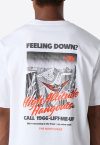 Osoba v bílém tričku s grafikou, na které je zobrazena osoba relaxující v houpací síti mezi horami, a textem propagujícím "Vysokohorské odpočinky."