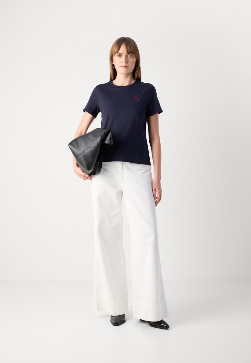 Navyblaues Baumwoll-T-Shirt mit Rundhalsausschnitt und kurzen Ärmeln, kombiniert mit weit geschnittenen weißen Hosen. Eine schwarze Ledertasche wird auf der Hüfte getragen.