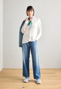 Woman wearing white blouse, wide-leg blue jeans, sneakers, colorful neck scarf, holding blue denim jacket over shoulder, standing on wooden floor.