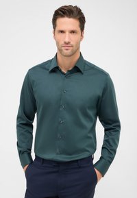 Camicia teal con colletto strutturato, maniche lunghe e bottoni scuri. Il tessuto ha una consistenza liscia ed è indossata con pantaloni scuri.