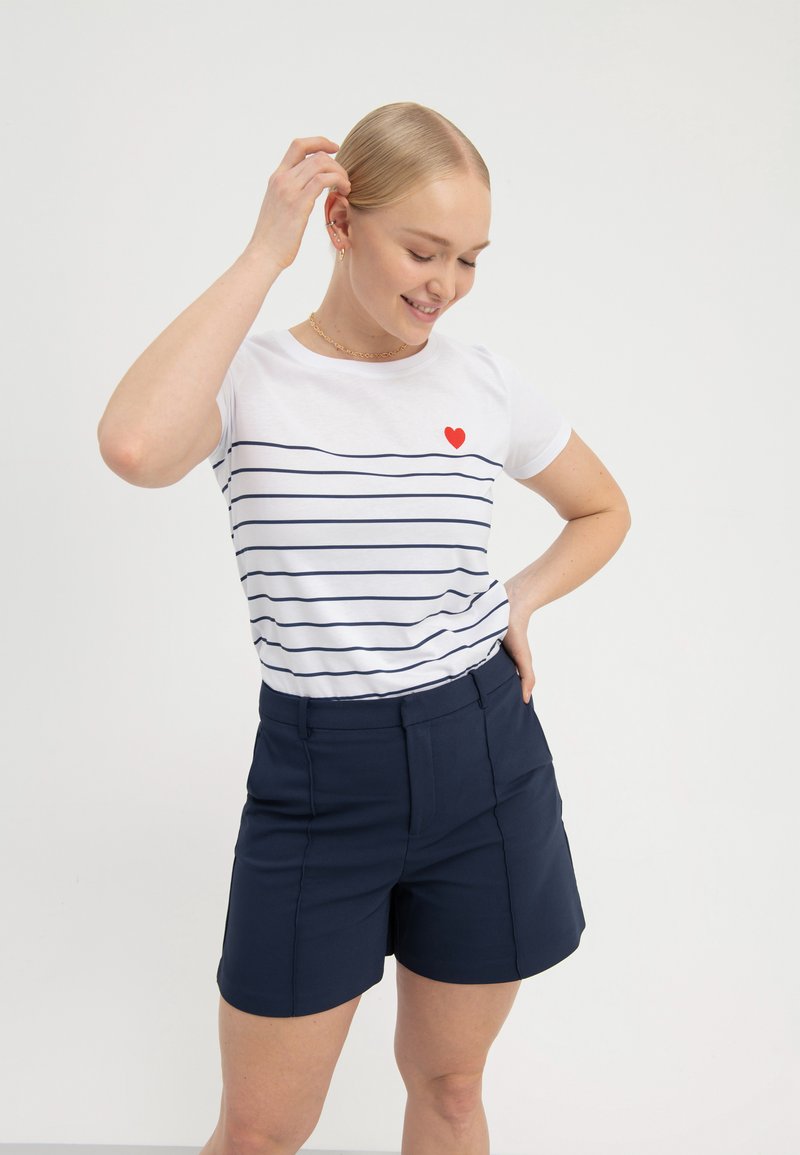 T-shirt bianco con righe orizzontali blu navy, accentuato da un piccolo cuore rosso. Abbinato a pantaloni corti blu navy scuro, caratterizzati da cuciture piatte e tasche.