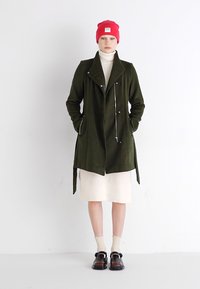 Abrigo verde oliva con cuello con muescas, cremallera frontal y bolsillos laterales, combinado con un vestido de punto crema, acompañado de zapatos negros y un gorro rojo.