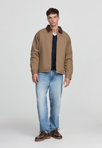 Giacca di stoffa marrone con colletto scuro, indossata sopra una camicia blu navy, abbinata a jeans azzurri chiari e scarpe marroni. Design semplice e casual.