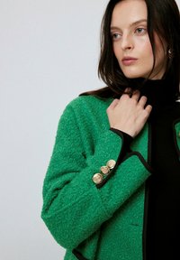 Sacou verde texturat cu detalii negre, având trei nasturi aurii la mansetă. Straturi de țesătură duale vizibile. Modelul poartă un pulover negru cu guler înalt.