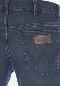 Jeans en denim bleu foncé avec un patch en cuir marron portant le logo Wrangler, featuring un design classique à cinq poches et une texture nervurée.