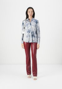 Camicia azzurro chiaro con stampa di palme, abbinata a pantaloni in pelle sintetica borgogna. Il modello indossa sandali bianchi con dettagli del logo.