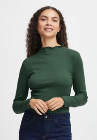 Långärmad, ribbad topp i mörkgrön med en vågig halsringning och manschetter, som bärs med mörkblå jeans. Slät textur med figurnära design.