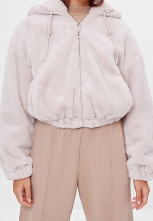 Nainen, jolla on cropped-tyylinen, vaalean beigen värinen pörröinen vetoketjuhuppari, jossa on nyörillinen huppu, ja yhteen sopivat, korkeavyötäröiset beigenväriset housut.