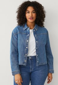 Giacca di jeans azzurra chiara con chiusura a bottoni, tasche frontali e colletto. Abbinata a una maglietta bianca e jeans blu, dal fit classico.