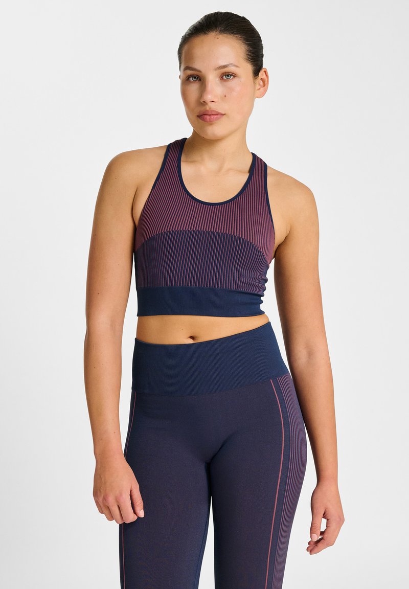 Frau trägt einen marineblauen und kastanienbraunen gestreiften sportlichen Croptop und hoch taillierte Leggings, steht vor einem schlichten weißen Hintergrund.