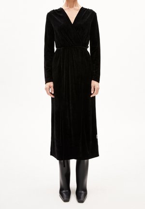 Femme portant une robe portefeuille longue en velours noire avec une ceinture à la taille, associée à des bottes noires montantes à bout carré, sur un fond blanc.