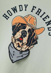 Šviesiai žalia marškinėlių su piešiniu, kuriame pavaizduotas Šv. Bernardo šuo, dėvintis oranžinį kaubojaus skrybėlę ir baltą, tarkuotą bandaną.