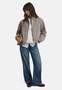 Woman wearing gray zip-up jacket, white cable knit sweater, striped shirt, wide-leg blue jeans, black shoes, and brown shoulder bag, standing.
