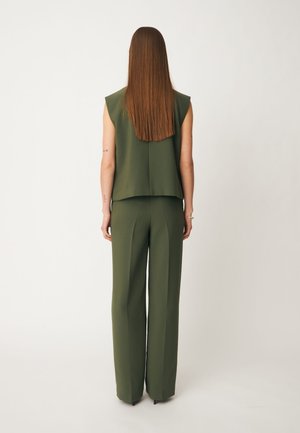 Femme aux longs cheveux raides, debout de dos, portant un top sans manches vert foncé et un pantalon large assorti sur fond uni.