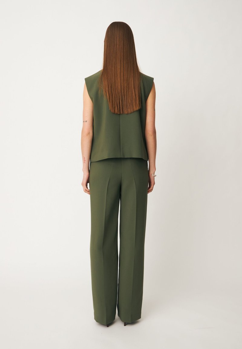 Femme aux longs cheveux raides, debout de dos, portant un top sans manches vert foncé et un pantalon large assorti sur fond uni.