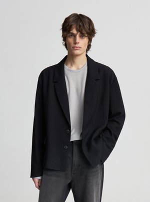 Giovane adulto con capelli castani di media lunghezza che indossa un blazer nero sopra una maglietta grigia e jeans grigio scuro, in piedi contro uno sfondo chiaro e semplice.