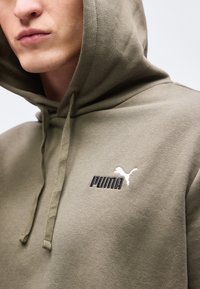 Κοντινό πλάνο ενός ατόμου που φοράει ένα ελιάς πράσινο φούτερ Puma με το λογότυπο της μάρκας στο στήθος και εμφανείς κορδόνια.