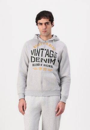 Grijze zip-up hoodie met een voorkPocket, voorzien van vetgedrukte tekst in zwart en oranje. Het materiaal lijkt zacht; heeft een capuchon en ribgebreide manchetten.