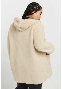 Beige fleecejacka med huva, med en strukturerad mjuk yta, oversize-passform och sidofickor, visad bakifrån.