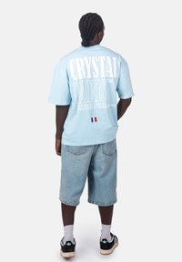 Světle modré oversized tričko s nápisem "CRYSTAL PARIS" v bílé barvě, v kombinaci se světle modrými denimovými šortkami a černými teniskami. Bílé akcenty a logo.