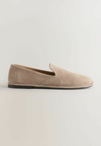 Chaussure beige en daim, modèle à enfiler, avec une semelle noire basse et des détails simples cousus, présentée de profil sur un fond uni.