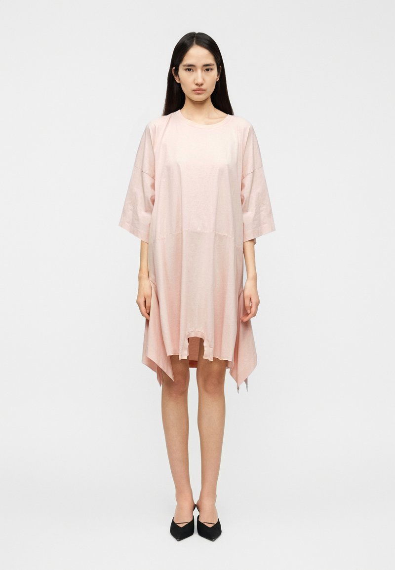 Robe ample rose clair à manches courtes, coupe ample, ourlet asymétrique et texture lisse. Portée avec des chaussures noires à bout pointu.