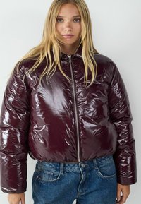 Burgunderfarbene Steppjacke mit glänzender Oberfläche, Reißverschluss, gerippten Bündchen und kurzer Passform, getragen über blauen Jeans.