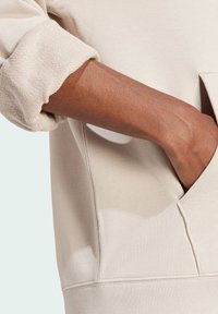 Beige sweatshirt with a textured, rolled cuff. Features a side pocket and subtle stitching detail. Fabric appears soft and comfortable.