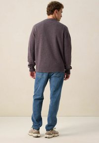 Pull en tricot gris avec des poignets et un ourlet côtelés, associé à un jean bleu clair. Le modèle se tient de dos, portant des baskets dans des tons neutres.