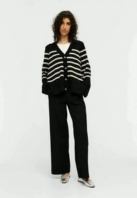 Cardigan à rayures noires et blanches avec un col V, fermeture à boutons et épaules tombantes, associé à un pantalon large noir et des ballerines argentées.