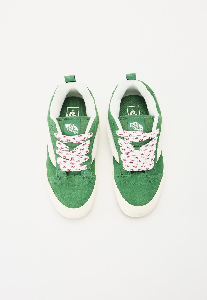 Baskets en suède vert avec des accents blancs, dotées de lacets à motifs floraux roses et blancs et d'un logo sur la languette, design minimaliste.