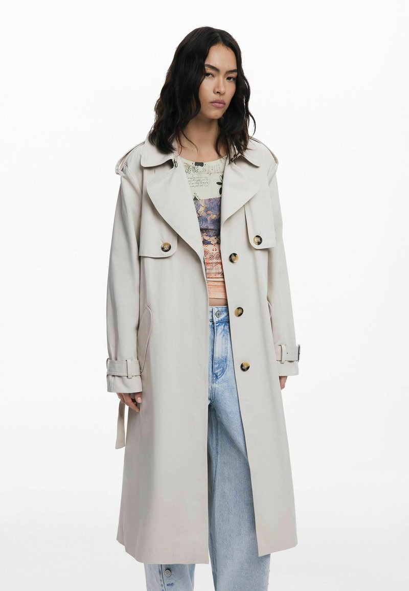 Beige trench coat with a structured design, large collar, gold buttons, tie waist, and side pockets, worn over a patterned top and wide jeans.