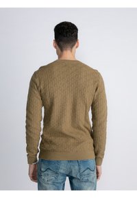 Khaki trui met een gestructureerd patroon, ronde hals en ribgebreide manchetten. G gedragen met lichtblauwe jeans, waarbij de achterkant zichtbaar is.