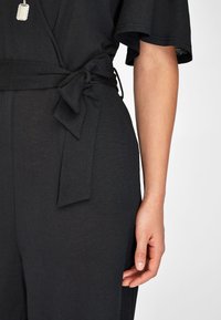 Tuta nera con tessuto texture, maniche larghe e design con vita legata. Vista ravvicinata della vita e della mano.