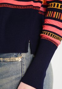 Gros plan sur une personne portant un pull rayé bleu marine et rose avec des coutures décoratives, associé à un jean denim bleu clair déchiré.