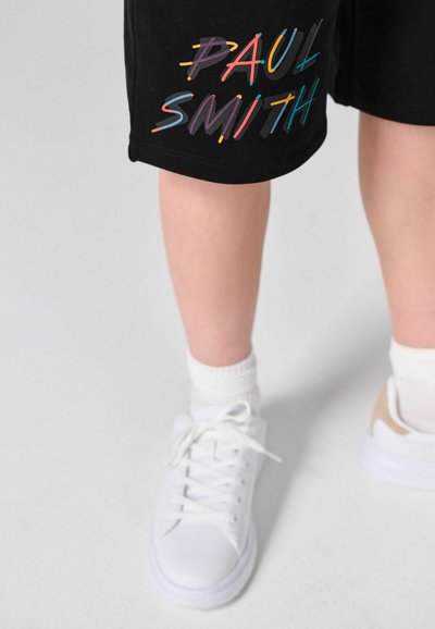 Jambes portant un short noir avec le texte coloré "Paul Smith", des chaussettes blanches à la cheville et des baskets blanches sur un sol gris clair.