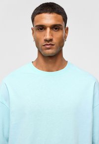 Sweat à col rond bleu clair en tissu doux, coupe décontractée avec épaules tombantes, présentant une texture lisse et des détails de couture minimalistes.
