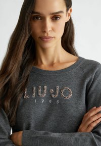 Felpa grigia con la scritta "LIU•JO 1995" embellita con borchie metalliche. Collo e polsini a costine, texture morbida e vestibilità rilassata.